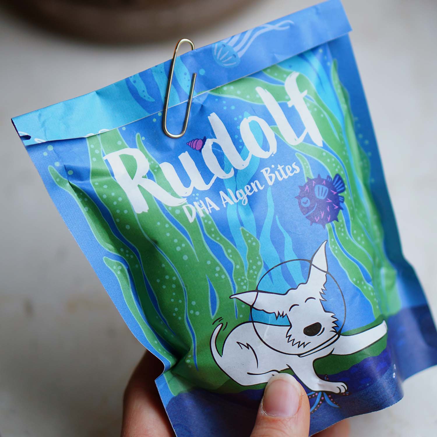 A hand holding the Vegan4Dogs Rudolf DHA Algae Bites pack, practically secured with a metal clip for daily DHA supply.
